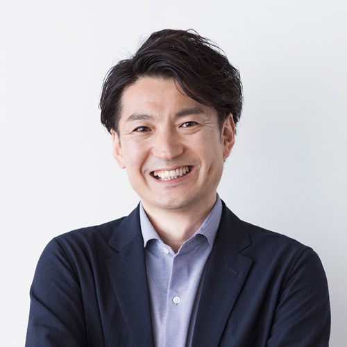 KDDIアジャイル開発センター株式会社 VPoE 兼 開発戦略本部 本部長 岡澤 克暢の笑顔のアップショット。紺色のスーツと空色のシャツを着ている。